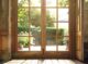 Photo d'une porte fenêtre en bois vernie, avec grain de bois visible, lumière naturelle traversant la porte. Une atmosphère paisible avec une vue sur un jardin.